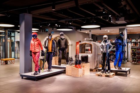 In einem modernen Geschäft wird stilvolle Skibekleidung ausgestellt. Puppen sind in farbenfrohen Skianzügen, Helmen und Schutzbrillen gekleidet. Im Hintergrund sind Regale mit Skiausrüstung und Zubehör zu sehen. Die Atmosphäre ist gemütlich und gut beleuchtet.