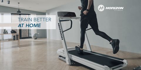 Persona che corre su un tapis roulant Horizon Fitness in uno spazio moderno e arioso con pavimenti in legno e ampie finestre. Accanto al logo di Horizon Fitness c'è il testo "Allenati meglio a casa".