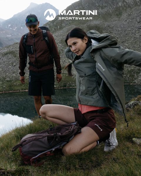 Due persone con abbigliamento da outdoor e zaini in spalla si trovano in riva a un lago di montagna. Una è inginocchiata e si sistema la giacca, l'altra è in piedi e sorride. Sullo sfondo, terreno roccioso e cielo nuvoloso. Nella parte superiore dell'immagine si legge "MARTINI SPORTSWEAR".