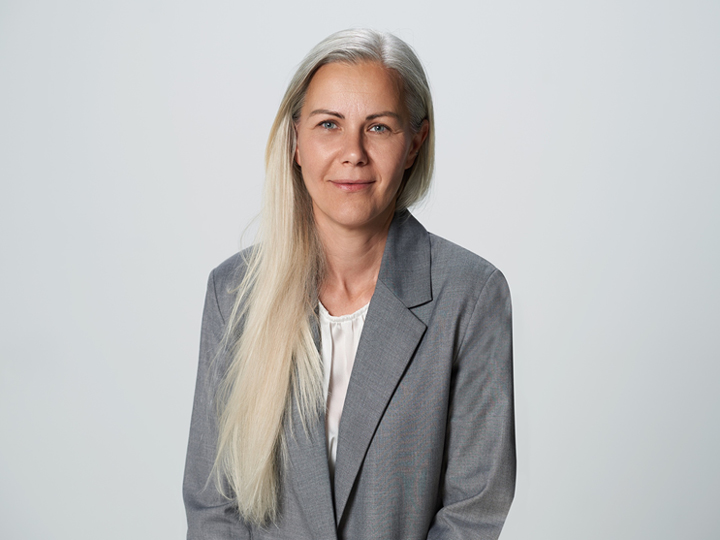 Una persona con capelli lunghi, lisci e chiari indossa un blazer grigio e una camicetta bianca. È seduta e guarda la fotocamera con un'espressione neutra su uno sfondo chiaro e semplice.