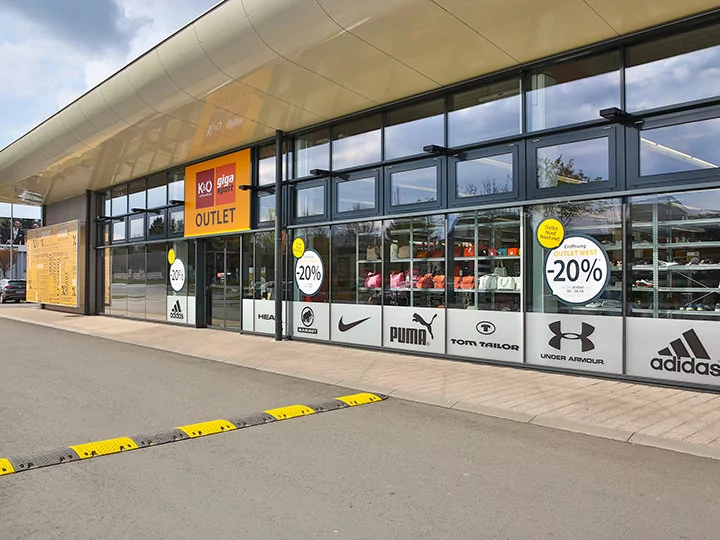 Vista esterna di un negozio outlet di articoli sportivi con ampie vetrine che mostrano vari loghi di marchi come Puma e Adidas. I cartelli sulle finestre pubblicizzano uno sconto del 20% su mobili, tende e tessili per la casa. L'edificio vanta un design moderno.