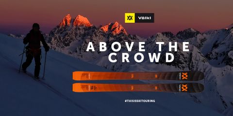 Uno sciatore si staglia come una silhouette contro il cielo al crepuscolo su una montagna innevata. Sullo sfondo si vedono le cime illuminate. In primo piano si vedono sci Völkl arancioni. Il testo recita "Above the Crowd" con gli hashtag #THISISSKTOURING.