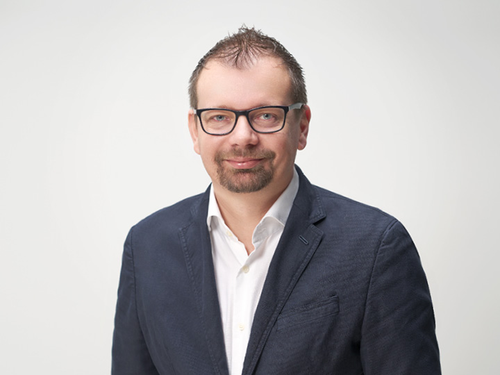 Un uomo con i capelli corti e gli occhiali indossa un blazer blu navy sopra una camicia bianca. Sorride e si trova di fronte a uno sfondo chiaro e semplice.