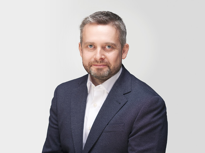 Un uomo con capelli grigi corti e barba indossa una giacca scura e una camicia bianca. È seduto di fronte a uno sfondo bianco semplice e guarda direttamente nella fotocamera con un'espressione neutra.