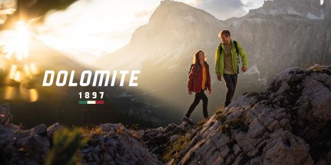 Un uomo e una donna con abbigliamento da trekking camminano su un terreno roccioso all'alba o al tramonto, con montagne sullo sfondo. Il testo recita "DOLOMITE 1897" con una piccola bandiera italiana sotto.