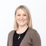 Una donna con capelli biondi di media lunghezza e un sorriso indossa un top nero e un cardigan marrone. Porta una collana di perle e si trova di fronte a uno sfondo bianco semplice.