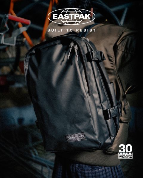 Una persona con una giacca marrone indossa uno zaino Eastpak nero con lo slogan "Built to Resist". L'immagine mostra un logo e la menzione di una "garanzia di 30 anni". Lo sfondo è sfocato.