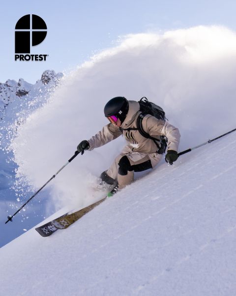 Uno sciatore con equipaggiamento beige e casco scia nella neve fresca su un pendio ripido, sollevando una nuvola di neve. Il logo "PROTEST" è visibile nell'angolo in alto a sinistra. Sullo sfondo si vedono un cielo limpido e montagne innevate.