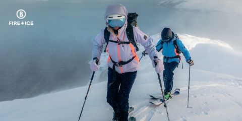 Due persone in abbigliamento invernale sciano su una montagna attraverso la neve. Indossano zaini e usano bastoncini per supporto. Sullo sfondo si vede un paesaggio nebbioso e vasto. L'immagine contiene un logo "Fire + Ice" nell'angolo in alto a sinistra.