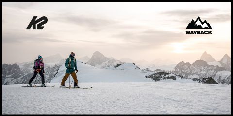 Due persone stanno salendo una montagna innevata sotto un cielo nuvoloso, con cime frastagliate sullo sfondo. I loghi di K2 e Wayback si trovano negli angoli superiori.