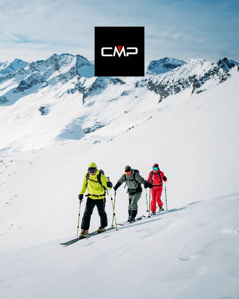 Tre sciatori in abiti colorati scalano una montagna innevata sotto un cielo azzurro e limpido, circondati da alte cime innevate. Il logo nero "CMP" appare in alto al centro dell'immagine.