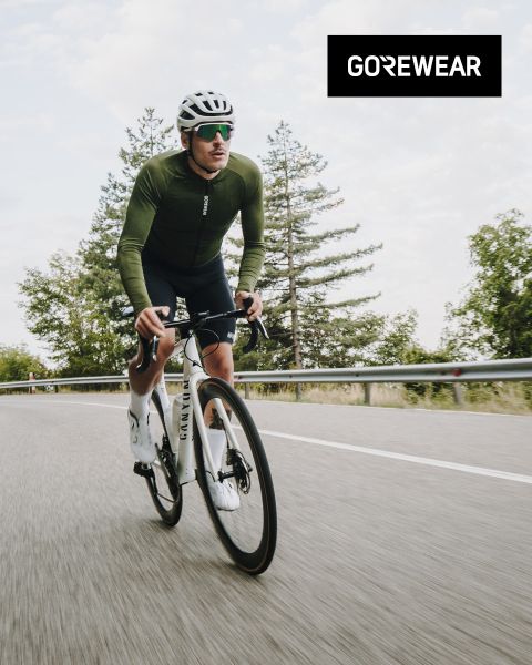 Ciclista con giacca verde e casco che pedala su una bicicletta bianca su una strada. Sullo sfondo si vedono alberi e un cielo nuvoloso. Il logo "GOREWEAR" è visibile in alto a destra.