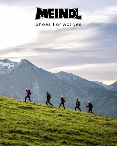 Un gruppo di escursionisti con zaini sale in fila una collina erbosa. Sullo sfondo si vedono montagne innevate e un cielo nuvoloso. Nella parte superiore dell'immagine si legge il testo "MEINDL Scarpe per persone attive".