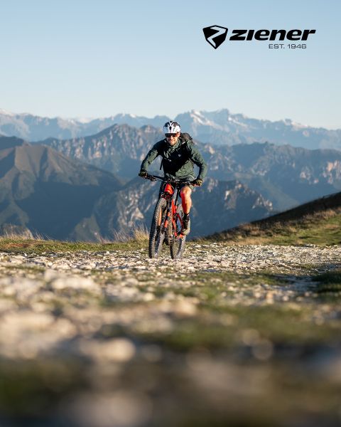 Un ciclista percorre un sentiero di ghiaia con la sua mountain bike rossa, sullo sfondo di aspre montagne. Il cielo è sereno, il ciclista indossa un casco e una giacca verde. In alto a destra si trova il logo Ziener.