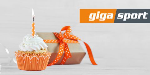 Ein Cupcake mit weißem Zuckerguss und einer orangefarbenen Kerze steht vor einem verpackten Geschenk mit einer orangefarbenen Schleife. Im Hintergrund ist das „Giga Sport“-Logo zu sehen.