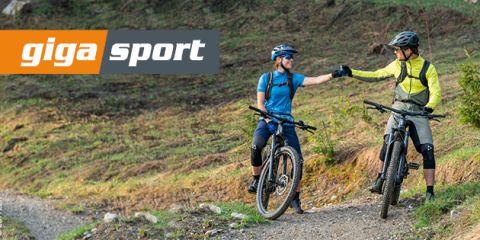 Zwei Radfahrer mit Helmen und Sportkleidung geben sich mit ihren Mountainbikes auf einem Grasweg einen Fauststoß. Oben links ist das Logo „giga sport“ zu sehen.