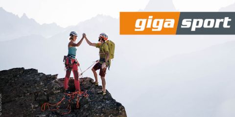 Zwei Kletterer mit Helmen und Ausrüstung geben sich auf einem Berggipfel vor nebliger Kulisse ein High-Five. Sie sind durch Kletterseile miteinander verbunden. Das Bild zeigt oben rechts ein großes „Giga Sport“-Logo.