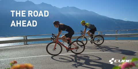 Due ciclisti su bici da corsa percorrono una strada di montagna tortuosa con vista su un pittoresco lago e montagne. Entrambi indossano caschi e abbigliamento da ciclismo. La luce del sole proietta ombre. Il testo sull'immagine recita "LA STRADA DAVANTI A NOI" con il logo Cube nell'angolo in basso a destra.