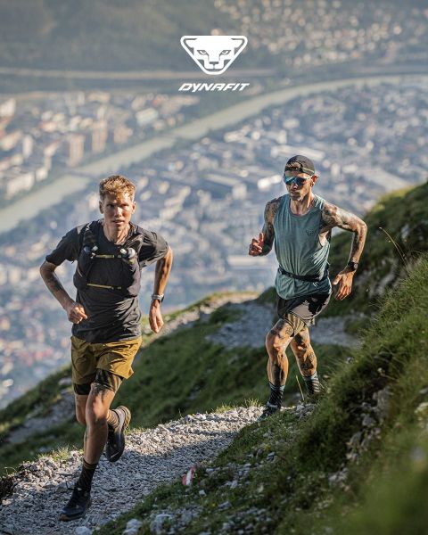 Due persone corrono su un sentiero di montagna con una città e un fiume sullo sfondo. Uno indossa una maglietta nera e pantaloncini, l'altro una canotta blu e pantaloncini. Il logo Dynafit si trova nella parte superiore dell'immagine.