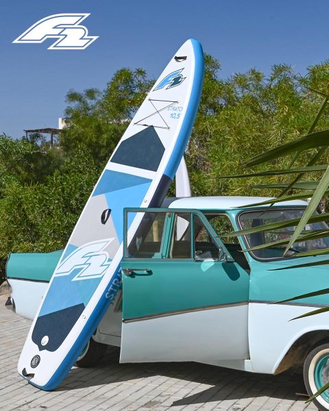 Un'auto d'epoca turchese e bianca è parcheggiata con una tavola da paddle blu e bianca appoggiata. Sullo sfondo si vedono alberi verdi e nell'angolo in alto a sinistra c'è un logo.