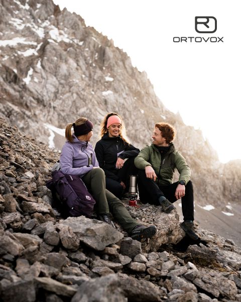 Tre persone con abbigliamento da outdoor sono sedute su un terreno roccioso in una regione montuosa, sorridendo e chiacchierando. Il sole tramonta dietro le cime. Uno zaino e un thermos sono nelle vicinanze. Il logo di Ortovox si trova in alto a destra.