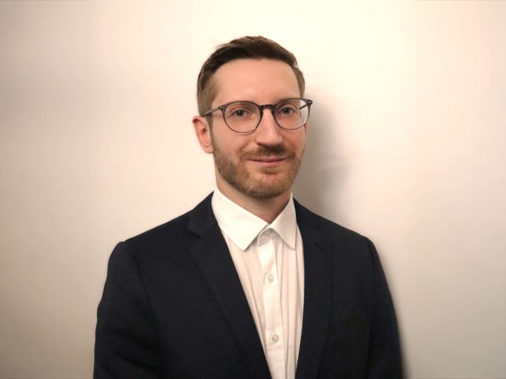 Una persona con occhiali, camicia bianca e blazer scuro è in piedi davanti a uno sfondo chiaro e semplice.