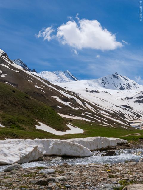 Un pittoresco paesaggio montano con cime innevate sotto un cielo azzurro e limpido. Un piccolo ruscello scorre in primo piano, fiancheggiato da campi di neve e da un pendio erboso. Sopra di esso fluttua una singola nuvola soffice.