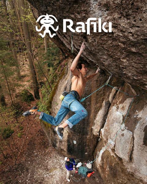 Un arrampicatore in jeans e imbracatura sta scalando una grande parete rocciosa a strapiombo, tenendosi alla roccia con una mano. Sotto, una persona in camicia bianca è in piedi a terra in mezzo alla sua attrezzatura da arrampicata. Nell'angolo in alto c'è la parola "Rafiki".