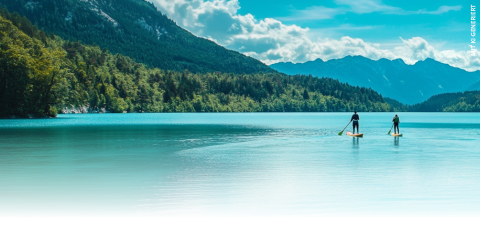 Due persone fanno stand-up paddleboarding su un tranquillo lago turchese, circondato da colline boscose e montagne in lontananza sotto un cielo azzurro con nuvole sparse.