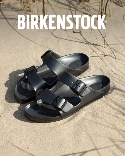 Una persona con sandali Birkenstock beige è in piedi accanto a un gioco d'acqua in un lussureggiante paesaggio di giardino. L'area è circondata dal verde e la parola "BIRKENSTOCK" è ben visibile sopra l'immagine.