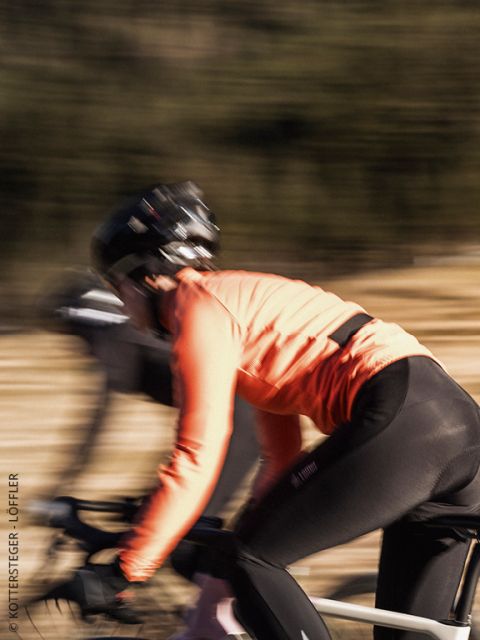 Un ciclista che indossa una giacca Löffler verde e un casco guarda avanti con uno sguardo concentrato. L'immagine è leggermente sfocata, il che enfatizza i dettagli della parte superiore del corpo e della giacca.