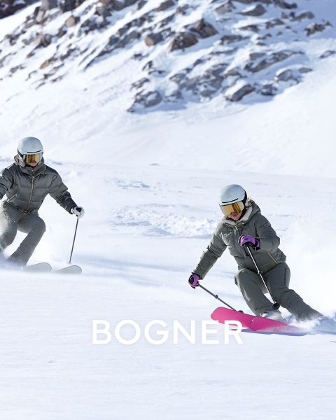 Due sciatori con attrezzatura colorata. A sinistra: persona in giallo con occhiali protettivi che tira una slitta nera. A destra: due persone in giacche gialle e blu che portano gli sci. Tutti indossano caschi e occhiali protettivi. Sfondo innevato con "BOGNER" nel testo.