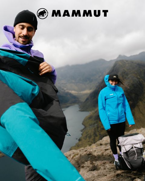 Due escursionisti con attrezzatura da outdoor attraversano un terreno roccioso sotto un cielo nuvoloso, con una grande montagna sullo sfondo. Entrambi indossano abbigliamento Mammut e il logo del marchio è visibile nella parte superiore dell'immagine.
