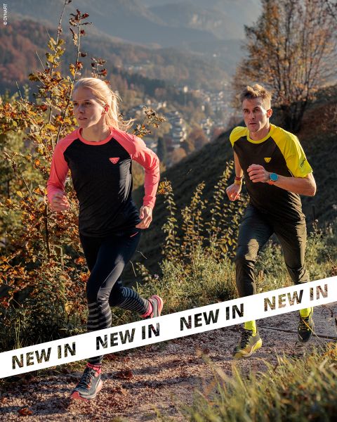 Due persone in abbigliamento sportivo corrono su un sentiero roccioso attraverso un paesaggio collinare e boscoso. La luce del sole illumina il fogliame autunnale. Banner bianchi con la scritta "NUOVO" in grassetto nero attraversano la parte inferiore dell'immagine.