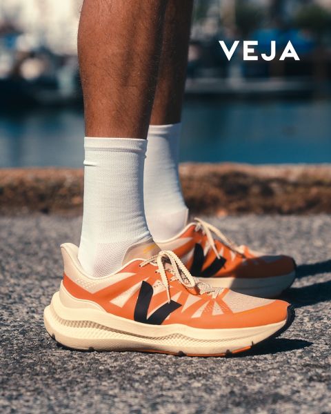 Una persona indossa sneakers Veja bianche con dettagli azzurri e suola in gomma. Le scarpe sono abbinate a pantaloni bianchi. Lo sfondo mostra un motivo geometrico su una parete piastrellata. Il logo Veja è visibile nell'angolo in basso a destra.