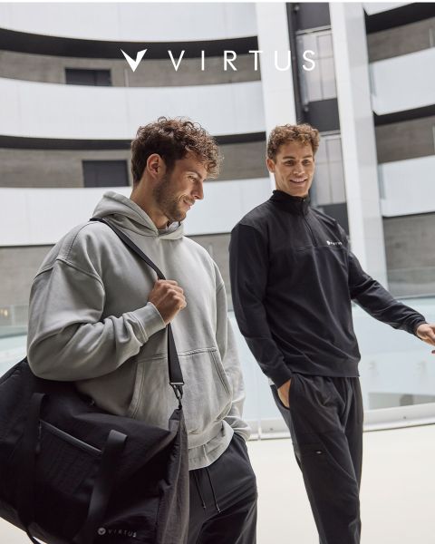 Due uomini in abbigliamento sportivo Virtus, uno con una felpa con cappuccio grigio chiaro e un borsone nero, l'altro con una giacca nera, sorridono e camminano insieme in una moderna hall. Il logo Virtus è visibile sopra di loro.
