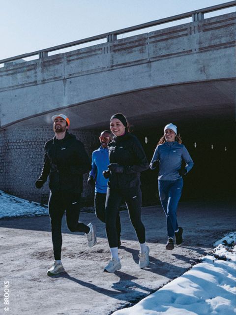 Quattro persone in abbigliamento da corsa invernale fanno jogging insieme su un sentiero innevato sotto un ponte di cemento, sorridendo e godendosi l'attività all'aperto.