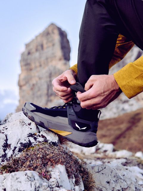 Una persona in abbigliamento da outdoor stringe i lacci di uno scarpone da trekking nero mentre si trova su una superficie rocciosa, con una grande formazione rocciosa e un cielo blu sullo sfondo.