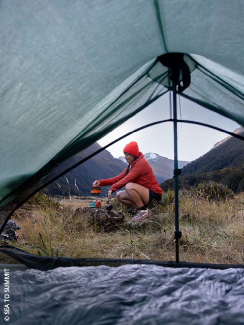 Una persona con giacca e cappello rossi prepara del cibo all'aperto, accovacciata nell'erba, vicino all'attrezzatura da campeggio, vista attraverso la porta aperta di una tenda con montagne e alberi sullo sfondo.