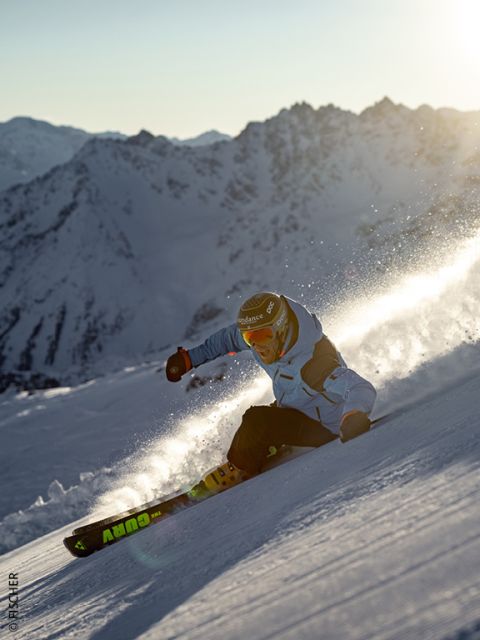Uno sciatore in giacca blu e con casco scende un pendio montuoso innevato al tramonto, sollevando neve fresca, con cime montuose aspre e illuminate dal sole sullo sfondo.