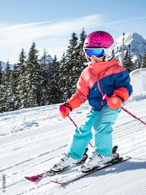 Un bambino piccolo con un casco rosa brillante, occhiali blu e un abbigliamento da sci colorato scende lungo una pista innevata, circondato da pini e montagne sotto un cielo azzurro limpido.