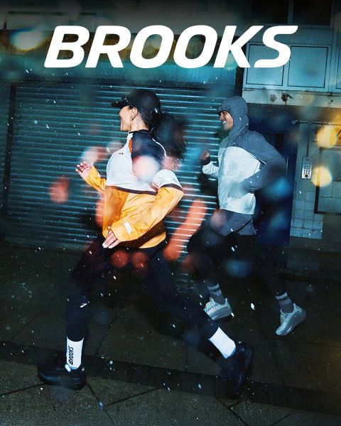 Un gruppo di cinque persone corre energicamente su una striscia pedonale. Indossano abbigliamento sportivo colorato, tra cui pantaloncini e canottiere. La scena è vivace, sopra di loro campeggia chiaramente la parola "BROOKS".