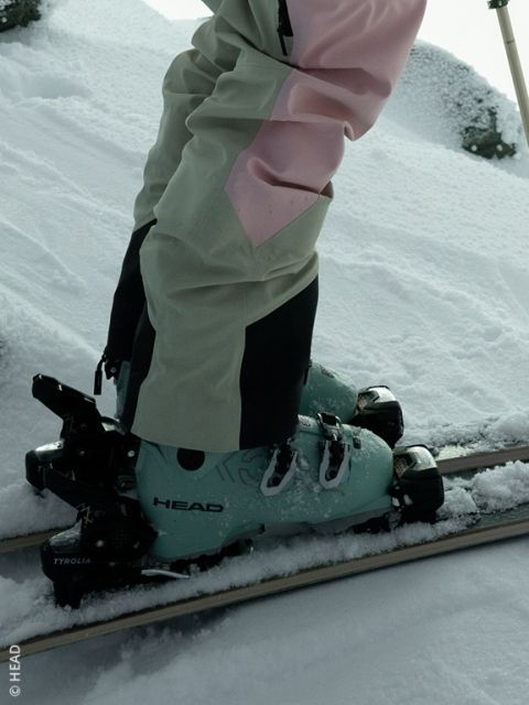 Una persona con scarponi da sci HEAD color verde menta e pantaloni da sci beige e rosa sta in piedi sugli sci nella neve fresca.