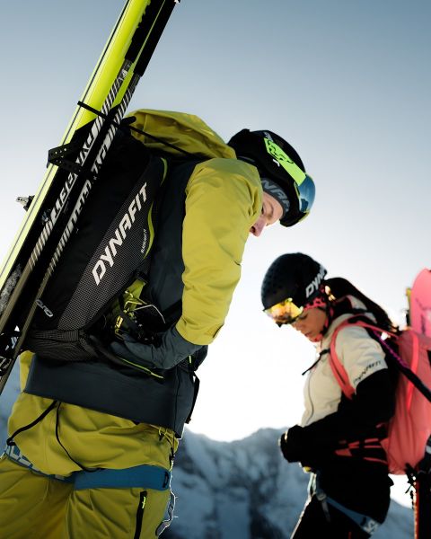 Due persone corrono su un sentiero di montagna con una città e un fiume sullo sfondo. Uno indossa una maglietta nera e pantaloncini, l'altro una canotta blu e pantaloncini. Il logo Dynafit si trova nella parte superiore dell'immagine.