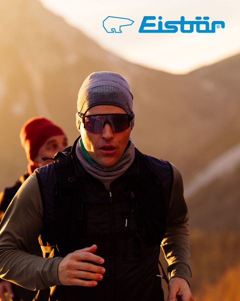 Una donna e un uomo sorridono mentre sciano in un paesaggio innevato. Lei indossa una giacca rossa e un cappello bianco e tiene in mano dei bastoncini da sci, lui una giacca beige e un cappello verde. Sopra di loro campeggia il logo "Eisbär".