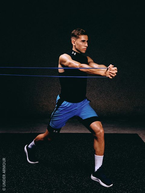 Un uomo in abbigliamento sportivo esegue un esercizio con elastici di resistenza, in posizione di affondo, allungando gli elastici con entrambe le braccia tese in avanti su uno sfondo scuro.