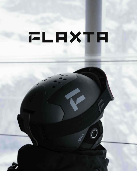 Due sciatori con caschi e attrezzatura si trovano su una piattaforma metallica innevata, con vista su un paesaggio montano innevato. Hanno gli sci in mano e sono uno di fronte all'altro. Sopra di loro, in grassetto, campeggia la parola "FLAXTA".