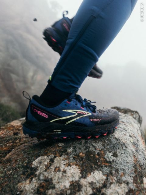Una persona con scarpe da trail running blu e leggings blu entra in un'area rocciosa all'aperto, con nebbia e rocce sullo sfondo.