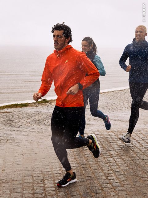 Tre persone fanno jogging sotto la pioggia su un sentiero di ciottoli bagnato vicino all'acqua. La persona davanti indossa una giacca rossa, mentre le due persone dietro indossano abiti più scuri. Intorno a loro si vede chiaramente la pioggia che cade.