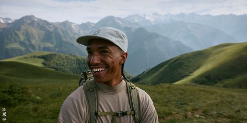 Una persona sorridente, che indossa un berretto leggero e uno zaino, si trova all'aperto in un paesaggio montano verde con cime lontane e nebbiose sotto un cielo parzialmente nuvoloso.